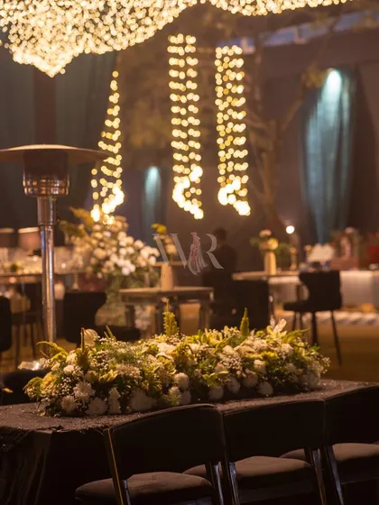 A simple yet elegant tablescape. A runner of white flowers sits on a black sequined tablecloth, with the soft, blurry lights of the fairy light canopy creating a beautiful bokeh effect in the background.
