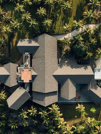 An aerial top-down view of a sprawling contemporary home in Mangalore. The complex roof geometry creates a dynamic form, while the layout is designed around a central pool and landscaped gardens, surrounded by a dense coconut grove.