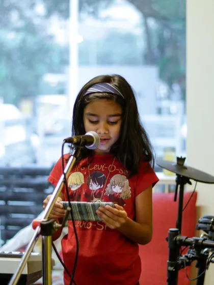 A young girl reading lyrics from her phone at the summer camp recital. We encourage them to perform songs they love.