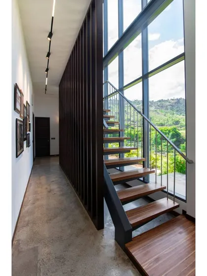 A minimalist staircase with wooden treads and a simple metal railing runs alongside a double-height window. This design choice ensures that the connection to the lush exterior landscape is never lost, even while moving between floors.