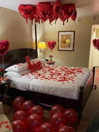 A classic romantic hotel room decoration with a bed covered in rose petals, a "Love" sign, and bouquets of red heart balloons.