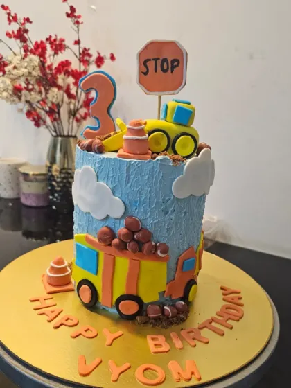 A tall construction themed cake for a 3rd birthday. The blue cake is decorated with fondant clouds, a dump truck, and a bulldozer, with crushed chocolate for dirt.