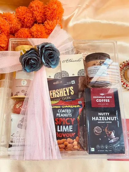 A clear acrylic box hamper filled with coffee and snacks, wrapped with a delicate pink ribbon and flowers.