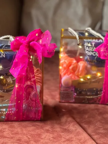 A pair of modern Rakhi hampers in clear boxes, lit up with fairy lights. These contain caramel popcorn, bracelets, and other fun items for a festive surprise.