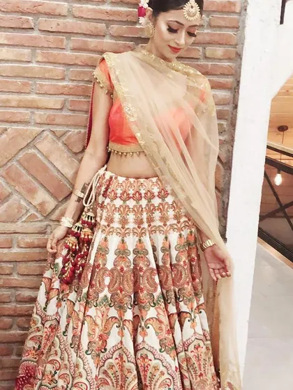 A client looking radiant in a designer lehenga by Rimple & Harpreet. The outfit features a vibrant printed skirt paired with a simple orange blouse and a light net dupatta, suitable for a mehendi or sangeet ceremony.