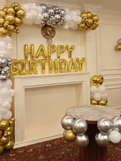 A sophisticated birthday setup in a living room with a fireplace. The decor features a gold, silver, and white balloon arch and 'Happy Birthday' foil letters.