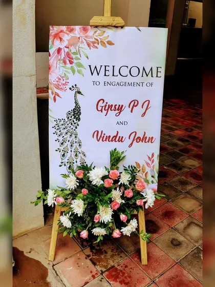 A beautifully designed welcome sign for an engagement, featuring an elegant peacock illustration. The sign is complemented by a lovely arrangement of pink and white flowers, adding a graceful touch.