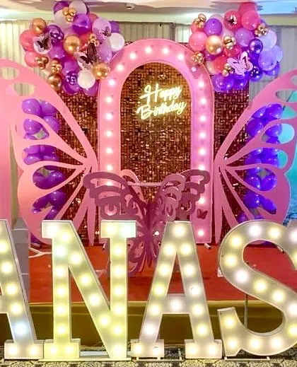 A close-up of a butterfly-themed stage with a pink sequin backdrop and large marquee letters. A custom photo of the birthday girl is integrated into the decor for a personal touch.