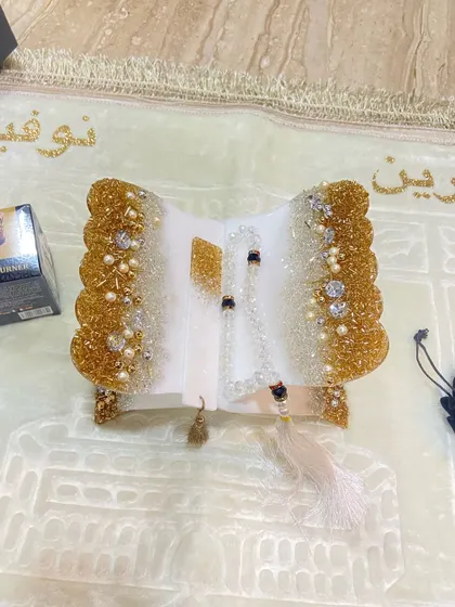 A white and gold Quran holder set, complete with a tasbih and bookmark, displayed on a personalized prayer mat.