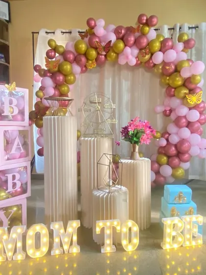 A 'Mom to Be' baby shower setup with a pink and gold balloon arch. The decor includes white plinths, 'BABY' blocks, and illuminated 'MOM TO BE' letters.