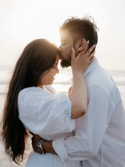 A tender moment as the groom-to-be kisses his fiancée's forehead on the beach at sunset. This is a classic, gentle, and romantic pose for any pre-wedding photoshoot.