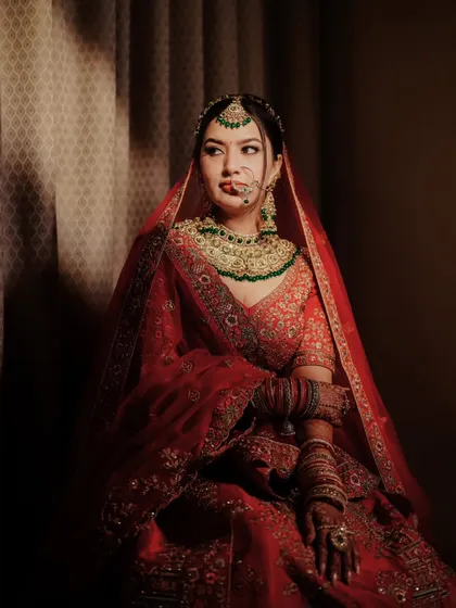 A thoughtful portrait of the bride, seated and looking away. The warm lighting enhances the rich red of her lehenga and her intricate jewelry.