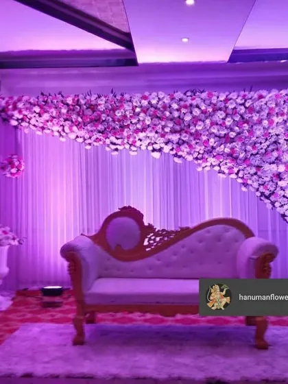 A beautiful stage designed for an engagement or reception. A classic white diwan is set against a backdrop of white curtains, framed by a cascading arch of pink and purple flowers, all under soft purple lighting.