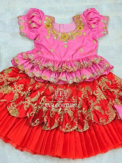A grand, layered pattu pavadai for a toddler. The pink peplum top is paired with a red skirt that has multiple layers and ruffles for a very festive look.