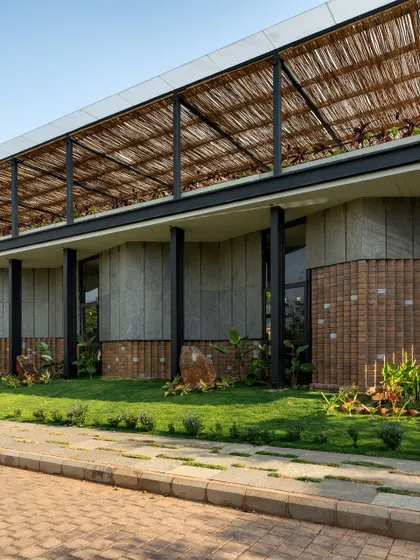 The building's facade features a combination of dry-clad granite and CSEB blocks, set against a lush lawn. The upper level is screened with a bamboo pergola, adding a layer of natural material that provides shade and texture.