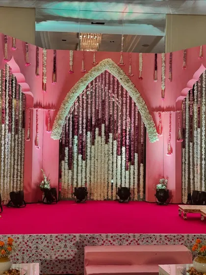 The main stage for a Bhaat ceremony. The backdrop features a pink wall with traditional Indian arches and cascading strings of white and pink flowers, creating a beautiful and respectful setting.