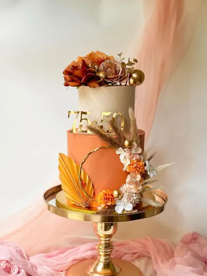 A stunning two-tier cake for a 75th birthday, featuring warm terracotta and cream tones. The design is accented with dried palm leaves, silk flowers, and gold details.