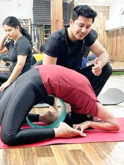 A teacher guides a student in Kapotasana (Pigeon Pose) using a yoga wheel, ensuring a safe and deep opening of the shoulders and thoracic spine.