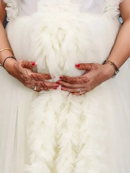 A close-up shot focusing on the details: henna-adorned hands with red nail polish gently holding a baby bump covered by a white ruffled gown. A beautiful detail of the pregnancy journey.