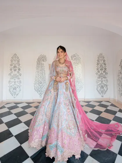The bride stands in a striking black and white checkered room, her elaborate pastel lehenga and pink veil creating a stunning visual.