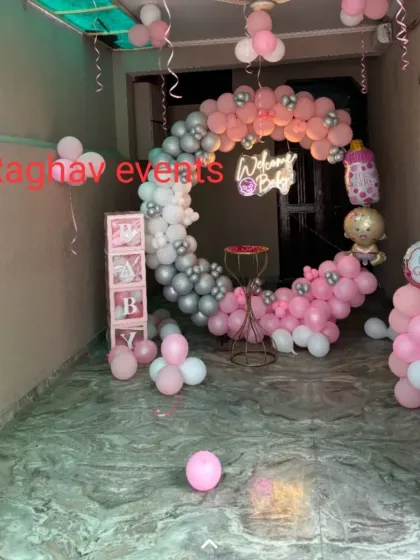 A wide view of a pink and silver themed welcome decoration for a baby girl. The setup includes a circular balloon arch, a neon sign, 'BABY' boxes, and other props.