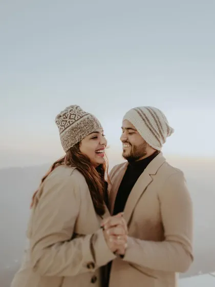 A happy couple, dressed in cozy winter wear and beanies, shares a laugh against the soft, hazy backdrop of the Himachal mountains. This candid moment captures the warmth of their connection in a cold climate.