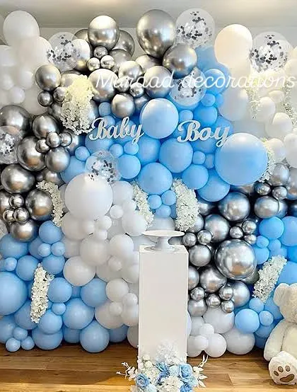 A classic 'Baby Boy' baby shower wall decoration. The dense balloon wall in shades of blue, silver chrome, and white creates a stunning backdrop, complete with a teddy bear and a pedestal for gifts or a cake.