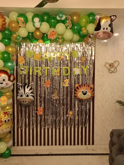 A fun jungle safari theme created indoors. This decoration uses a silver fringe backdrop framed by a green, gold, and white balloon arch, complete with various animal-head foil balloons like a lion, monkey, and giraffe.