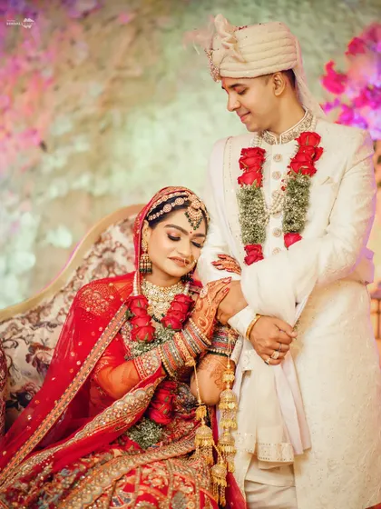 A tender moment where the groom holds his bride's hand. Her traditional red lehenga and his white sherwani create a classic and beautiful color contrast.