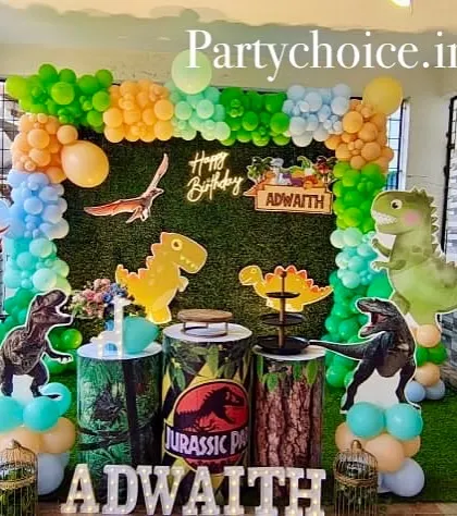 A dinosaur-themed birthday party setup in an outdoor covered area. It features a green wall backdrop, various dinosaur cutouts, and Jurassic Park-themed plinths.