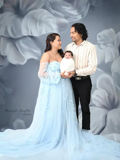 A classic family portrait with a newborn. The parents stand together holding their baby, dressed in a beautiful blue gown that matches the artistic floral backdrop.