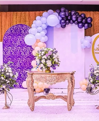 A full view of the royal prince stage setup. The design features multiple backdrops in purple and white, an ornate golden table, and elegant floral stands, all tied together with balloon arches.