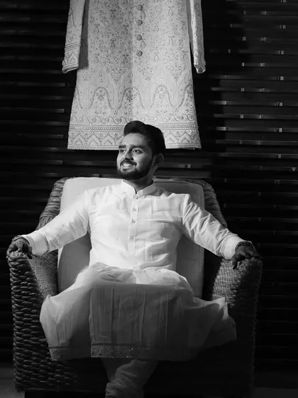 A black and white portrait that emphasizes the texture and silhouette of the groom's outfit. The sherwani hangs in the background, showing the full ensemble before the ceremony.
