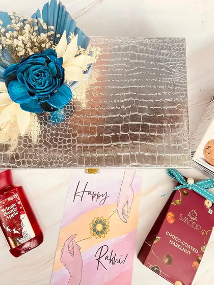 A stylish Rakhi gift set with a silver croc-textured box, containing cookies, chocolates, and a scented lotion.