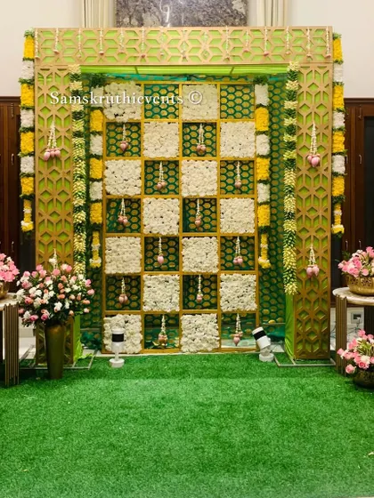 A modern and stylish backdrop featuring a green and gold jaali frame. The center is a checkerboard of white flowers, with hanging lotus buds adding a delicate, traditional touch.