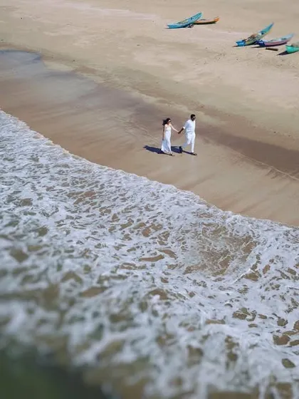 A high-angle drone shot captures a couple in white outfits standing at the water's edge on a wide, sandy beach. This image shows the grand scale of the location, making the couple's moment feel both epic and private.