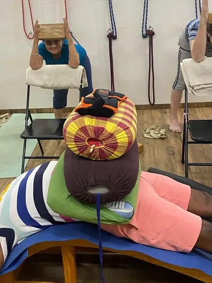 A student in a restorative pose with the legs supported and weighted. In the background, other students are in a supported forward bend, showing the variety of our practice.
