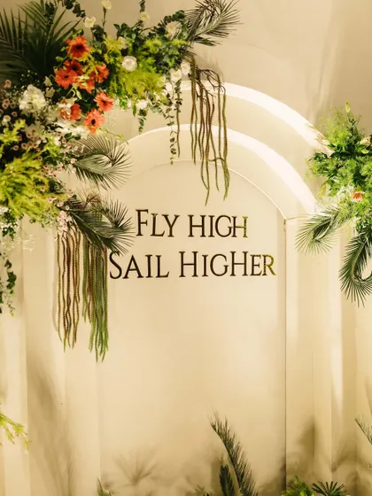 This custom arched backdrop for a joint 50th and 80th birthday features a personal quote, "Fly High Sail Higher," framed by lush tropical greenery and florals, making it a meaningful centerpiece.