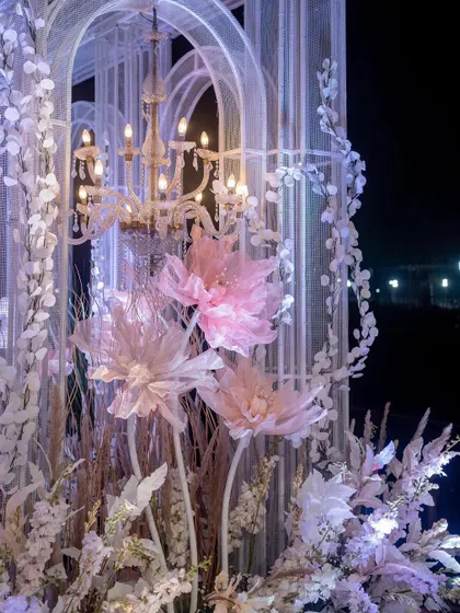 Details make the design. Here, oversized pink paper flowers are nestled within the grand, white architectural frames, lit by the warm glow of a vintage-style chandelier, adding a touch of whimsical artistry.