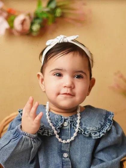 A close-up of a sweet baby girl, dressed in a denim shirt and pearls, giving a little wave. Her personality shines in this adorable portrait.