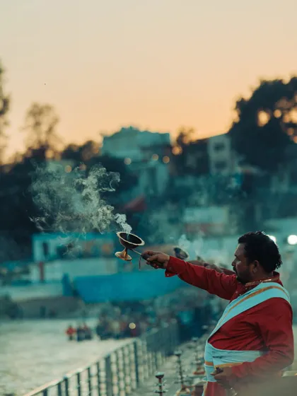 A priest holds an incense burner during the Ganga Aarti at sunset in Rishikesh. The soft evening light adds a warm, peaceful glow to the scene.