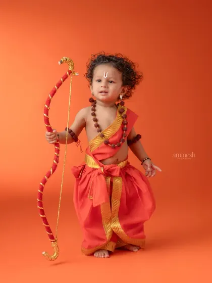 A simple and striking portrait of a baby dressed as Rama against a solid orange background. This minimalist setup makes the costume and the child's pose stand out.