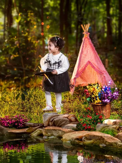 A toddler girl with a toy crossbow stands by a small pond in a whimsical outdoor setting. This imaginative photo combines nature with a sense of adventure.