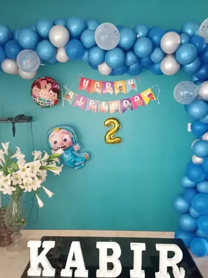 A simple Cocomelon decoration for a home setting. A blue and white balloon garland frames a 'Happy Birthday' banner and character foil balloons.