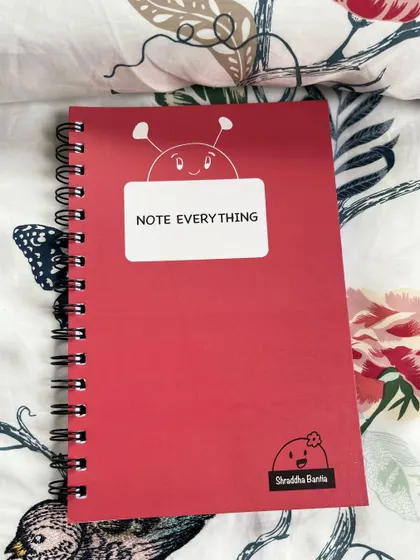 The red A5 notebook, with a cute bug doodle peeking over the "Note Everything" label.