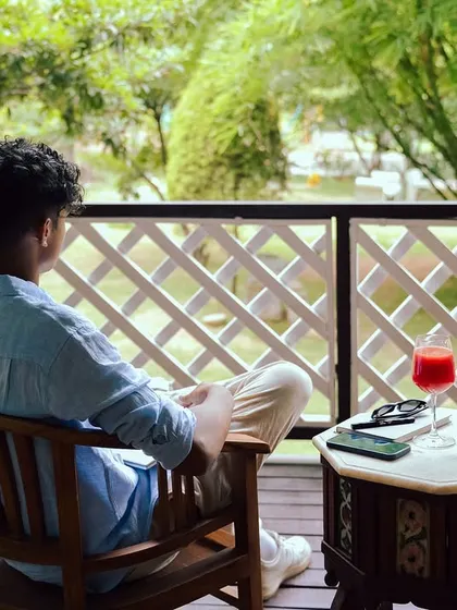 A guest takes a moment to soak in the peaceful evening vibes from a balcony chair, enjoying the lush green view and a cool drink.