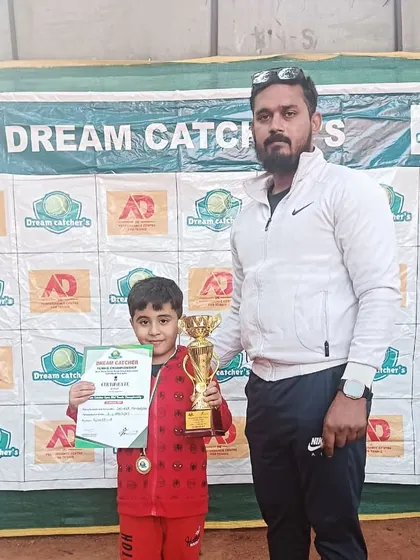 A proud coach with one of our youngest champions in the U6 category. The player holds his winner's trophy and certificate in front of the Dream Catcher's tournament banner.