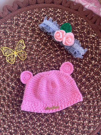 This image shows the pink bear-ear beanie next to a delicate crochet flower headband. Both are handmade and make a beautiful gift set for a baby.