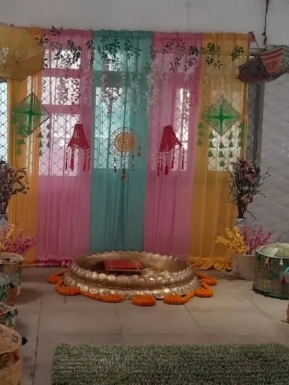 An indoor Haldi setup with a backdrop of colorful sheer curtains against a window. The space is furnished with traditional 'mooda' seating and a central 'urli' for the ceremony.