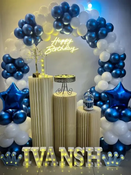 A stylish birthday party with a blue and white balloon arch, plinths, and star-shaped foil balloons. The name 'Ivansh' is spelled out in marquee letters.